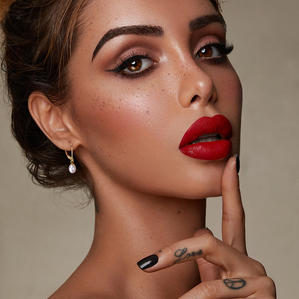 Porträt einer Frau mit roten Lippen, Perlenohring, dunklen Augenbrauen und Fingertattoo mit den Worten "Love" und einem Peace-Zeichen
