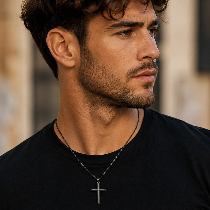Portraet einer Person mit schwarzem T-Shirt und dunkler Halskette mit Kreuzanhaenger, Blick zur Seite vor unscharfem hellem Hintergrund.